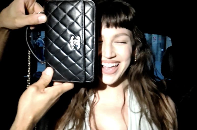 Woman smiles beside a black Chanel wallet with silver hardware, held by a person, in a dimly lit indoor environment.