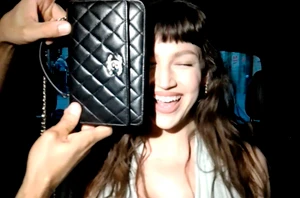 Ursula Corbero smiling and laughing while holding a black quilted Chanel handbag up to cover part of her face.