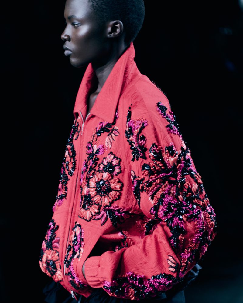 Runway fashion show featuring a black female model wearing a pink floral embellished jacket against a dark background in a studio setting.
