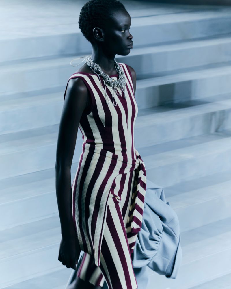 Runway model in striped dress and necklace on grey steps, captured in medium shot for fashion content with neutral color grading.
