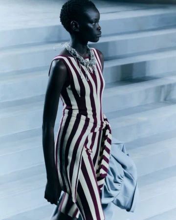 Runway model in striped dress and necklace on grey steps, captured in medium shot for fashion content with neutral color grading.