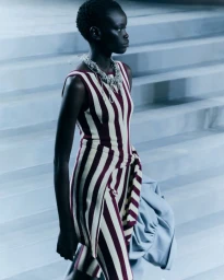 Black female model wearing a burgundy and cream striped sleeveless dress and silver necklace on pale steps.