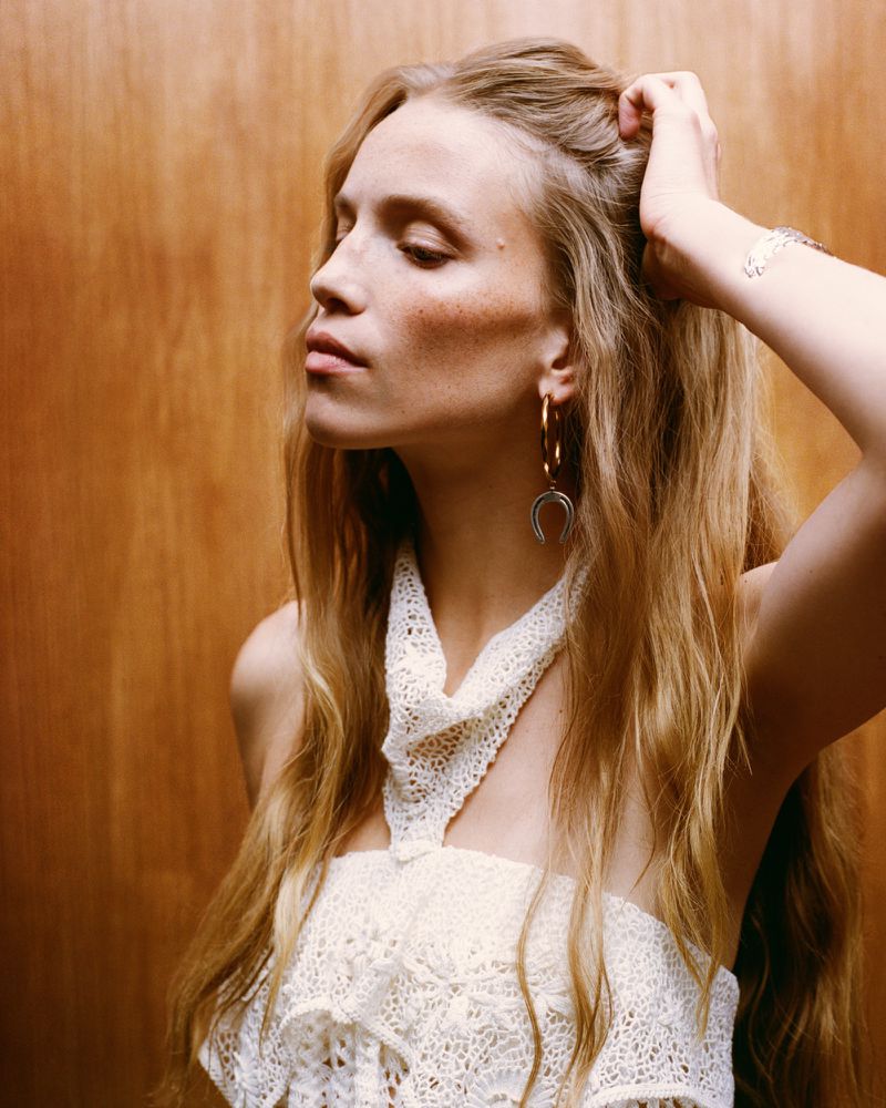Blonde woman poses in a close-up studio shot, wearing a lace top and hoop earrings, set against a neutral wood background.