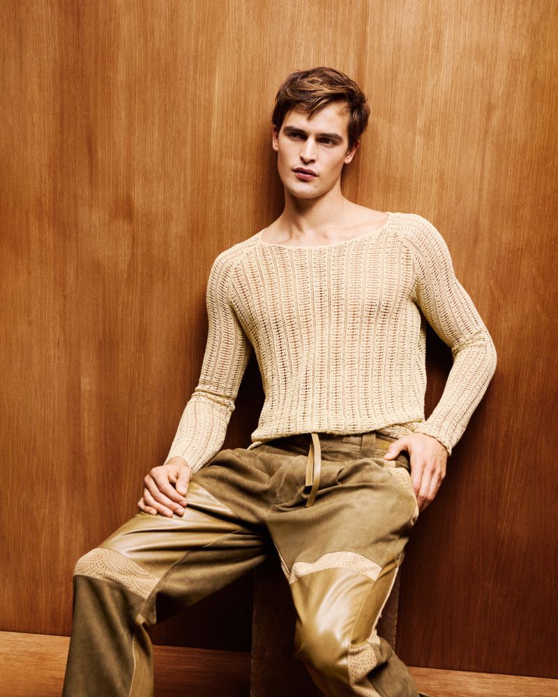 A fair-skinned adult male model poses indoors in a cream-colored knitted sweater and brown leather-patched pants against a wood background.