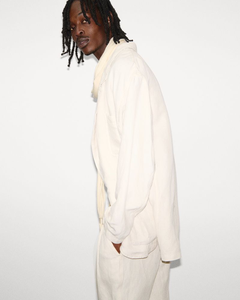 Portrait of a young adult male in a light beige suit standing against a white background, captured in a studio setting with soft lighting.