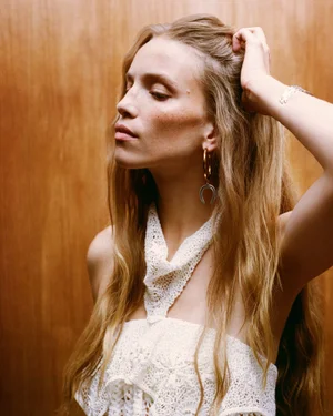 Model with long blonde hair wearing a white crochet halter top posing against a warm wood paneled wall.