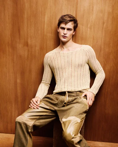 Male model seated against a wood-paneled wall wearing a cream knit sweater and olive trousers.