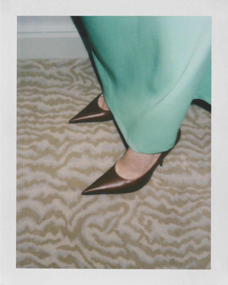 Close-up of a woman's legs in a pair of brown pointed-toe kitten heels, standing on a carpet with a light brown wave pattern. 