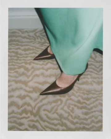 Close-up of a woman's legs in a pair of brown pointed-toe kitten heels, standing on a carpet with a light brown wave pattern. 