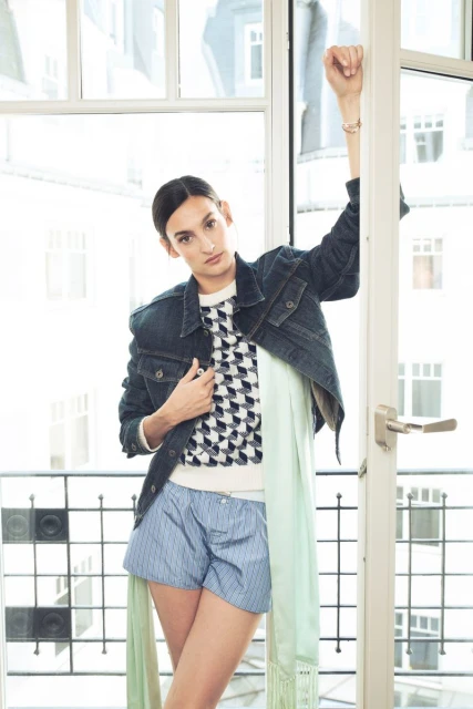 A woman poses in front of a window wearing a denim jacket, patterned sweater, and boxer shorts in an indoor setting.