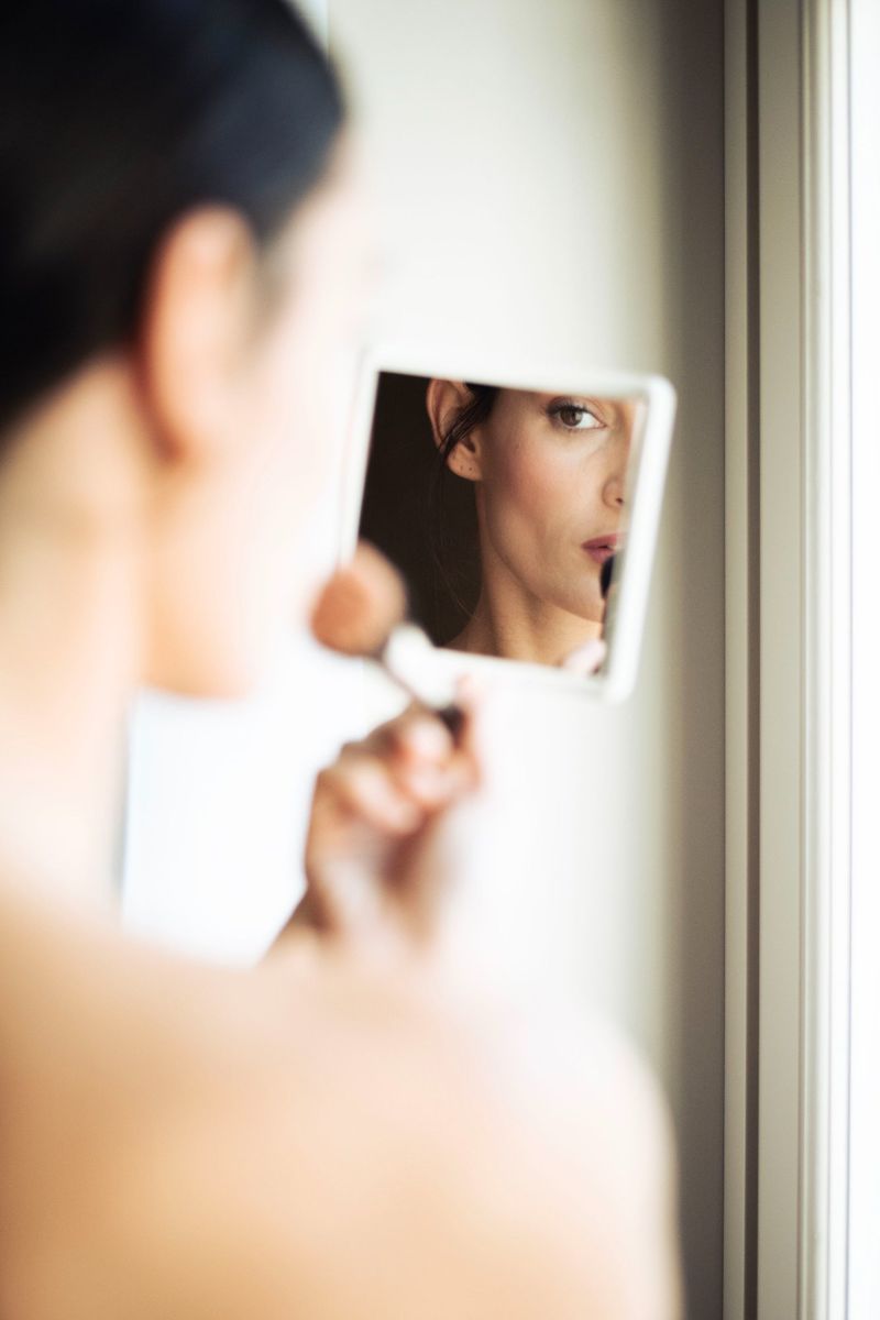 Woman applies makeup while looking in a hand mirror by the window in an indoor setting with soft lighting, creating a beauty moment.