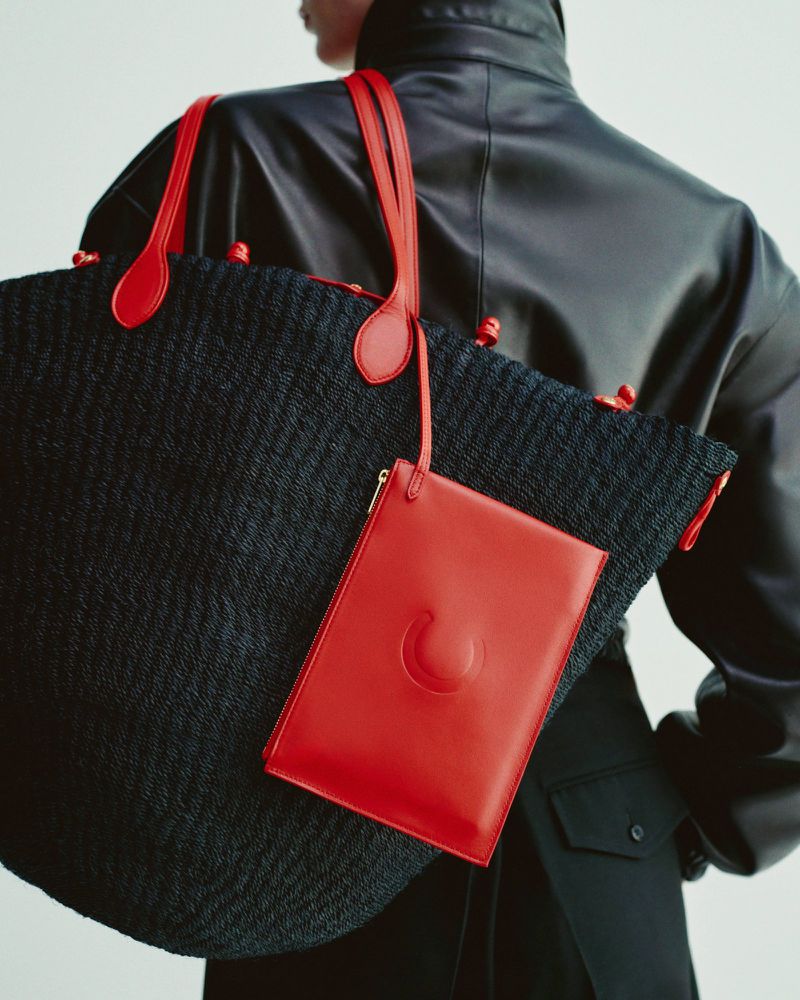 Black woven tote bag with red handles and a detachable red pouch, carried over the shoulder of a person wearing a black jacket.