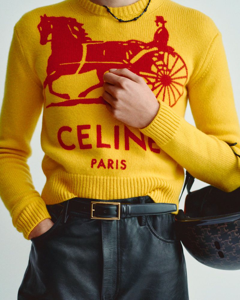 Person wearing a yellow CELINE sweater with a red horse-drawn carriage graphic, black leather pants and belt, holding a helmet.