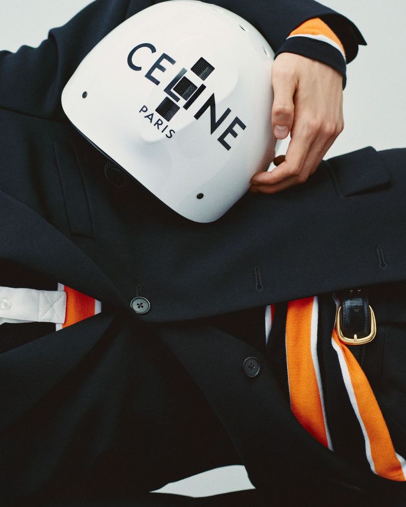 Close-up of a person wearing a dark blazer holding a white helmet with a bold logo. The image has a fashion editorial style.