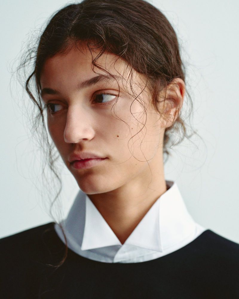 Portrait of a young woman with dark curly hair, wearing a black top and a white collared shirt, looking off to the side in a studio setting.