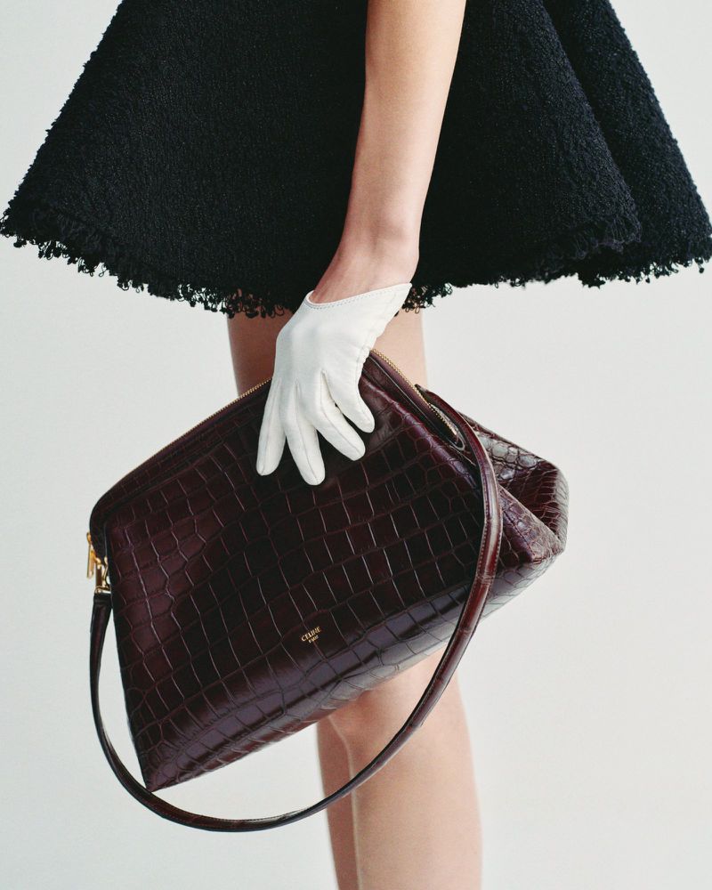 A model showcases a structured, dark brown crocodile-leather handbag with a top handle, paired with white gloves and a short black dress.