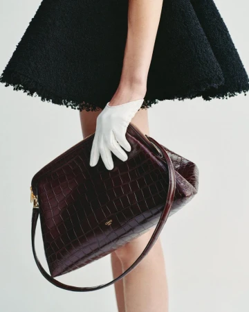 Model wearing a black skirt holding a dark red crocodile leather handbag with a white leather glove against a plain white background.