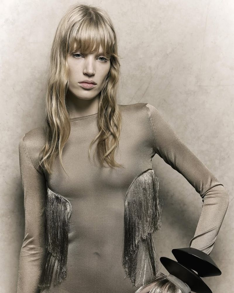 A fair-skinned fashion model with long wavy blonde hair poses indoors wearing a long-sleeved dress with fringe detailing and black wristbands.
