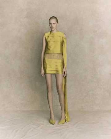 A single fashion model poses in a chartreuse-colored mini dress and matching heels against a light neutral mottled studio backdrop.