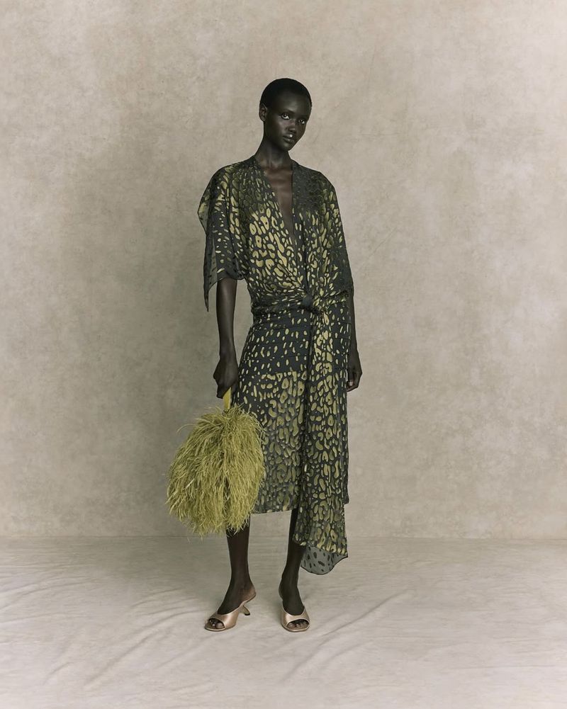 Woman in flowing green and black animal print dress poses in studio holding fluffy purse, elegant and fashionable.