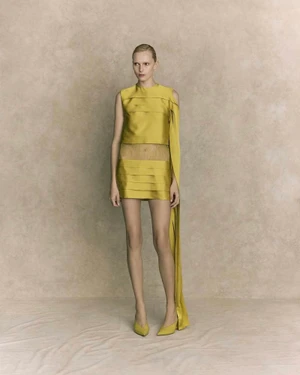 Model wearing a chartreuse pleated mini dress and matching heels against a neutral textured studio backdrop.