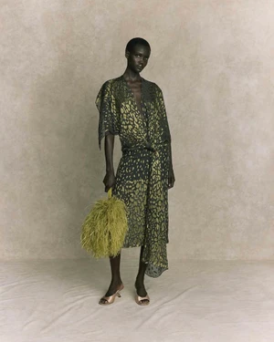 Model in printed sheer dress holding a textured green bag against a neutral background