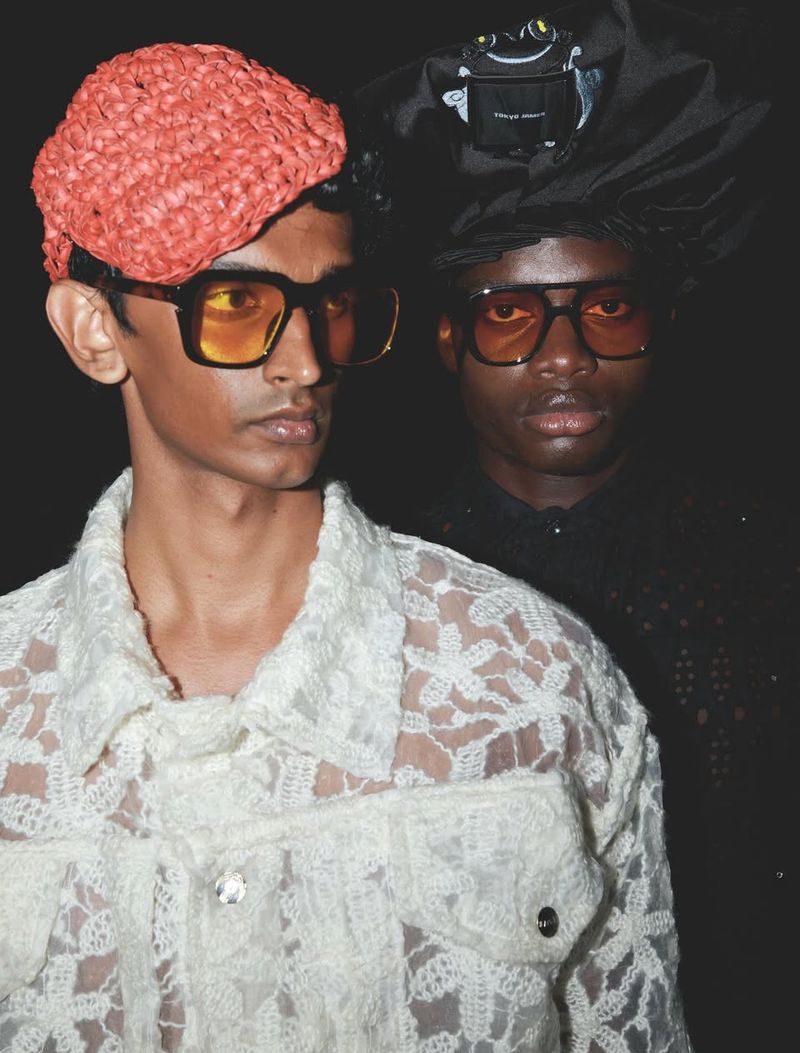 Two fashion models pose in studio lighting. One wears a straw hat and lace jacket, the other a black hood with frog detail.