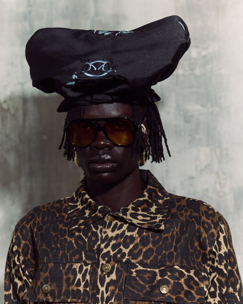 Portrait of an adult female model in a leopard print jacket and large sunglasses, wearing an unusual black hat on a grey background.