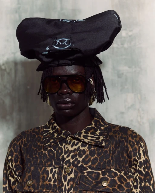 Portrait of an adult female model in a leopard print jacket and large sunglasses, wearing an unusual black hat on a grey background.