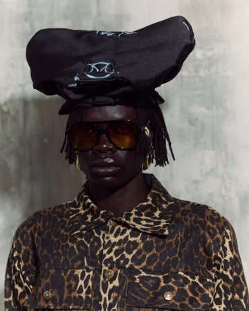 Portrait of an adult female model in a leopard print jacket and large sunglasses, wearing an unusual black hat on a grey background.