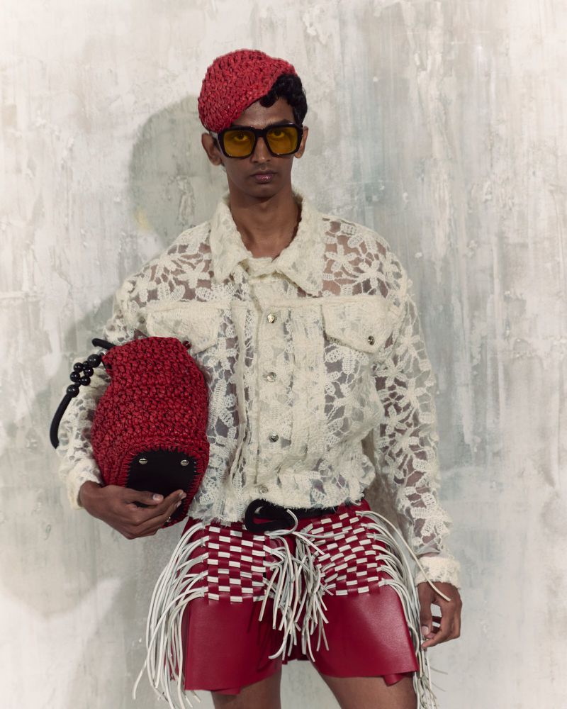 An adult male poses in a studio wearing a lace shirt, red shorts with fringe, a red beret, and yellow-tinted sunglasses, holding a red textured bag.