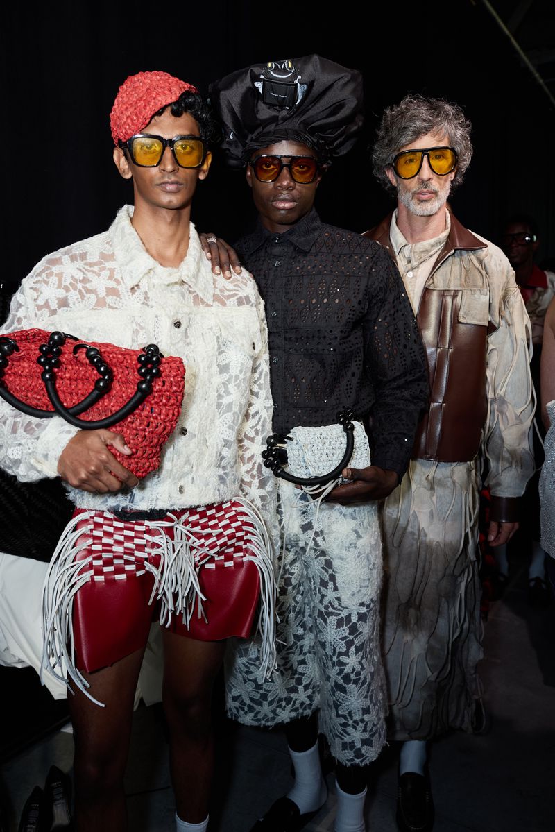 Three fashion models pose indoors against a dark backdrop, showcasing designer apparel and handbags in a portrait-style fashion shot.