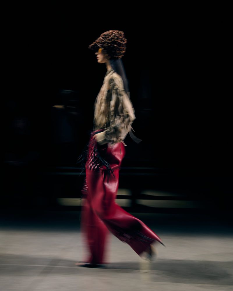 A blurred, full shot of a fashion model walking the runway in an avant-garde outfit including a hat and red wide-leg leather pants.