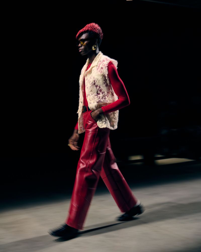 Runway fashion: a model walks in bright red leather pants and a white crochet vest against a black backdrop, showcasing a bold look.