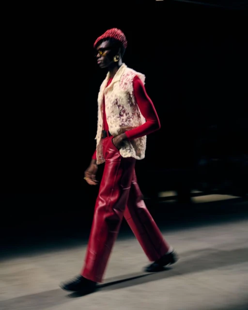 Runway fashion: a model walks in bright red leather pants and a white crochet vest against a black backdrop, showcasing a bold look.