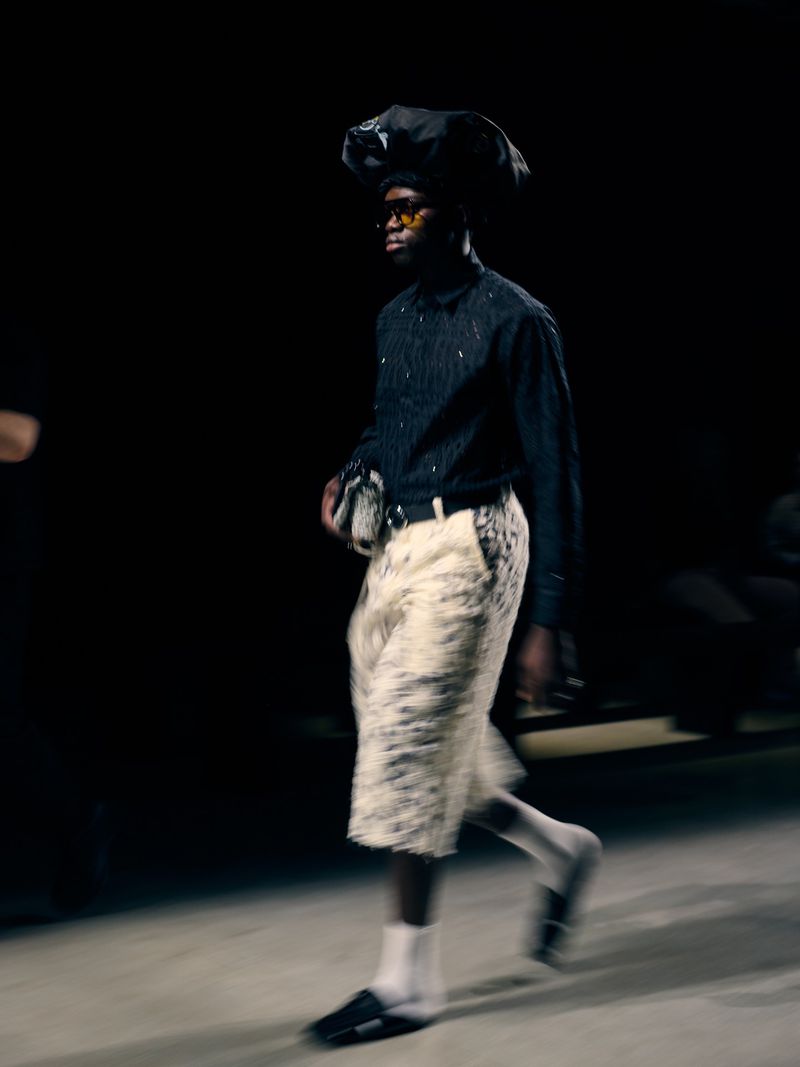Runway model walks in a fashion show wearing a dark shirt and white shorts, featuring a blurred motion effect.