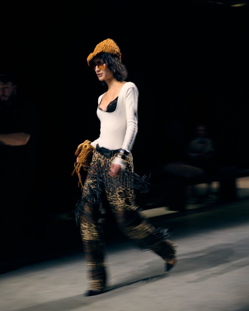 Runway model in patterned outfit and knit cap walks during a fashion show, captured in motion with blurred background and stage lighting.