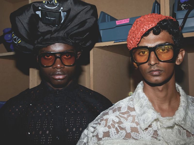 Two stylish men wearing sunglasses, hats, and patterned tops pose indoors against shelves, showcasing a fashion-forward editorial look.