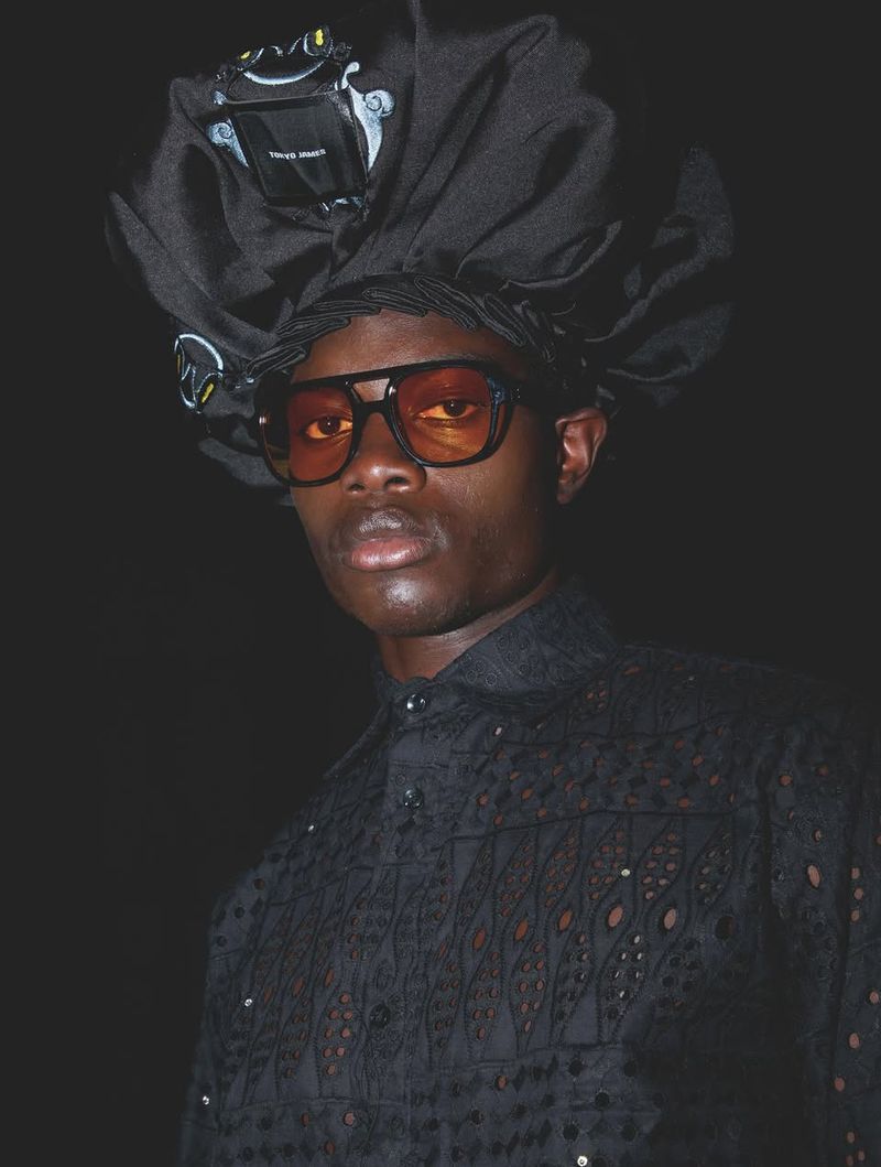 Close-up of a Black male model wearing orange tinted aviator sunglasses and an ornate black headpiece and shirt against a dark background.