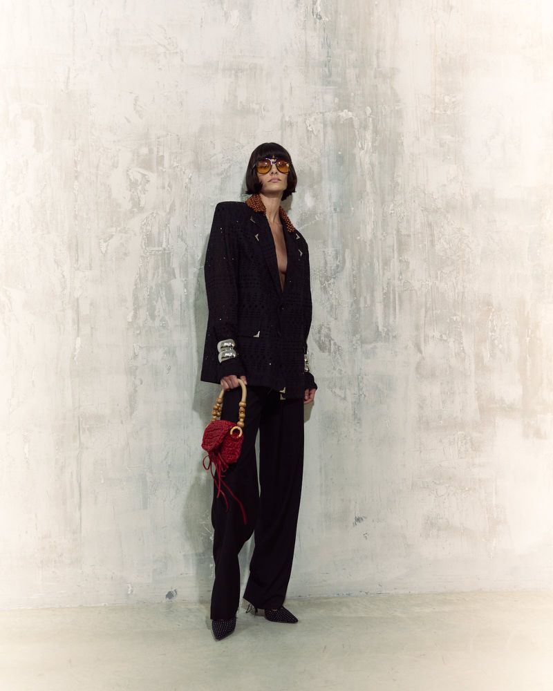 Woman poses in a black blazer and pants with a red purse against a textured wall, highlighted by indoor lighting. Fashion shot.
