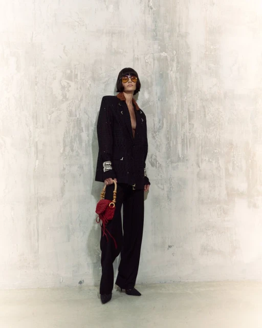 Woman poses in a black blazer and pants with a red purse against a textured wall, highlighted by indoor lighting. Fashion shot.