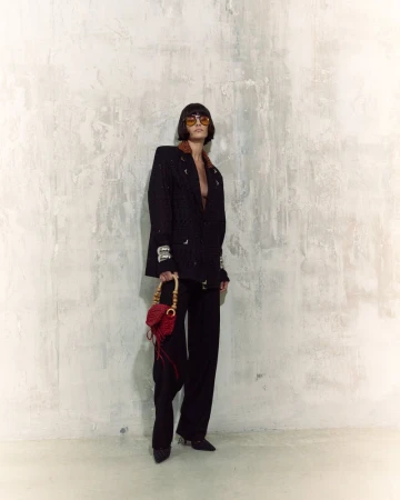 Woman poses in a black blazer and pants with a red purse against a textured wall, highlighted by indoor lighting. Fashion shot.
