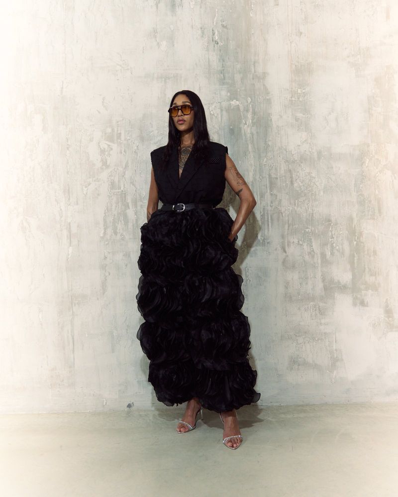 Woman in a sleeveless black top and large black ruffled gown posing against a textured white wall wearing sunglasses and heels.