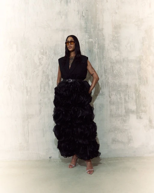 Woman in a sleeveless black top and large black ruffled gown posing against a textured white wall wearing sunglasses and heels.