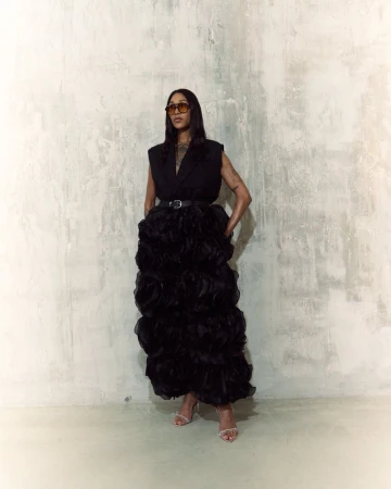 Woman in a sleeveless black top and large black ruffled gown posing against a textured white wall wearing sunglasses and heels.