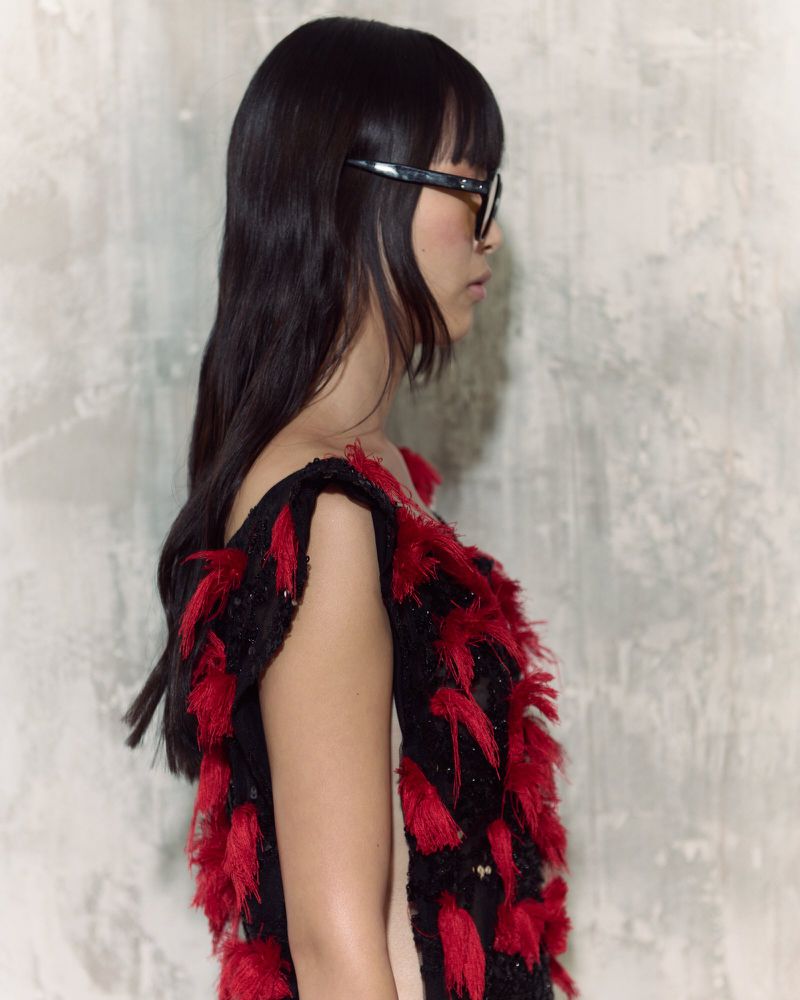 Side view of a woman with dark hair and glasses wearing a black dress with red fringe against a neutral textured background in soft light.