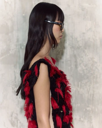 Side view of a woman with dark hair and glasses wearing a black dress with red fringe against a neutral textured background in soft light.