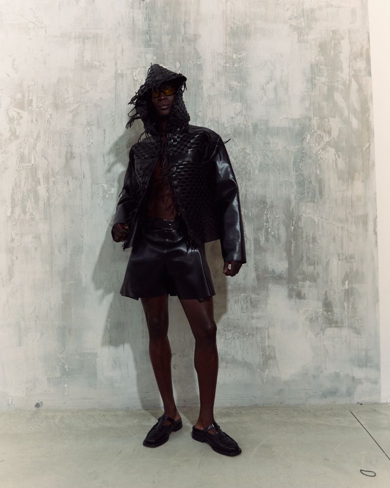 Dark-skinned adult wearing a woven hooded jacket and shorts, posing indoors against a distressed gray wall in a fashion shot.