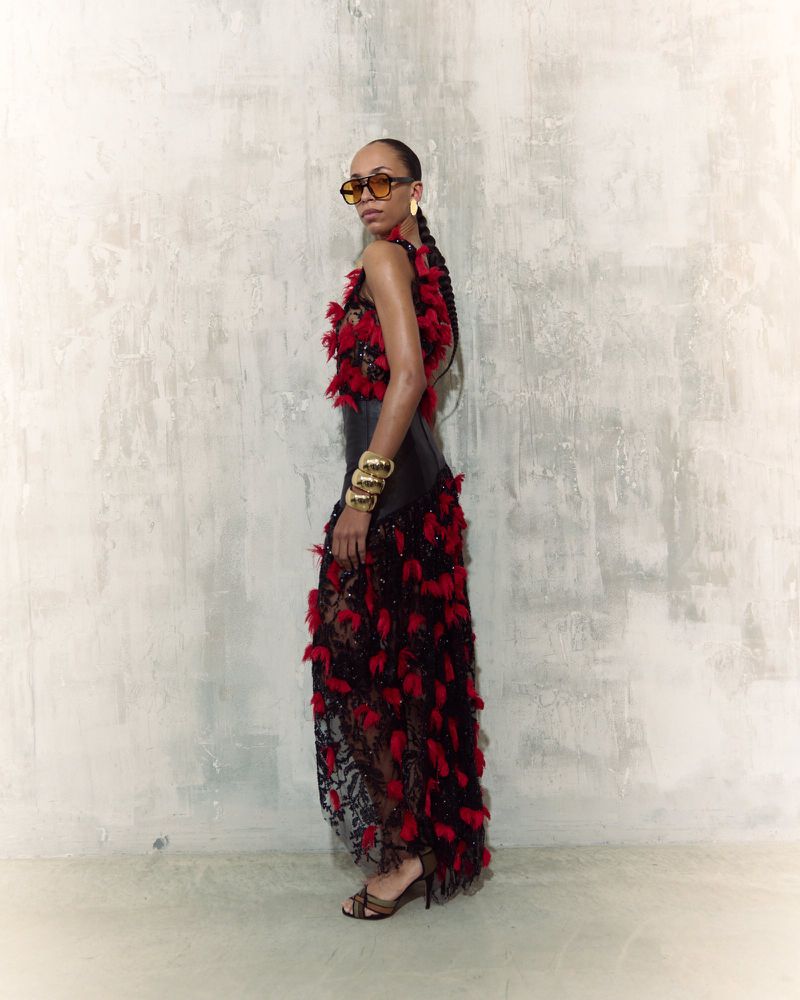 Elegant woman poses in a black and red feather dress against a textured wall. She wears sunglasses and gold bracelets.