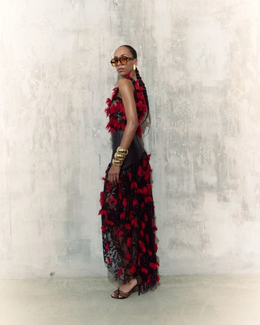 Elegant woman poses in a black and red feather dress against a textured wall. She wears sunglasses and gold bracelets.